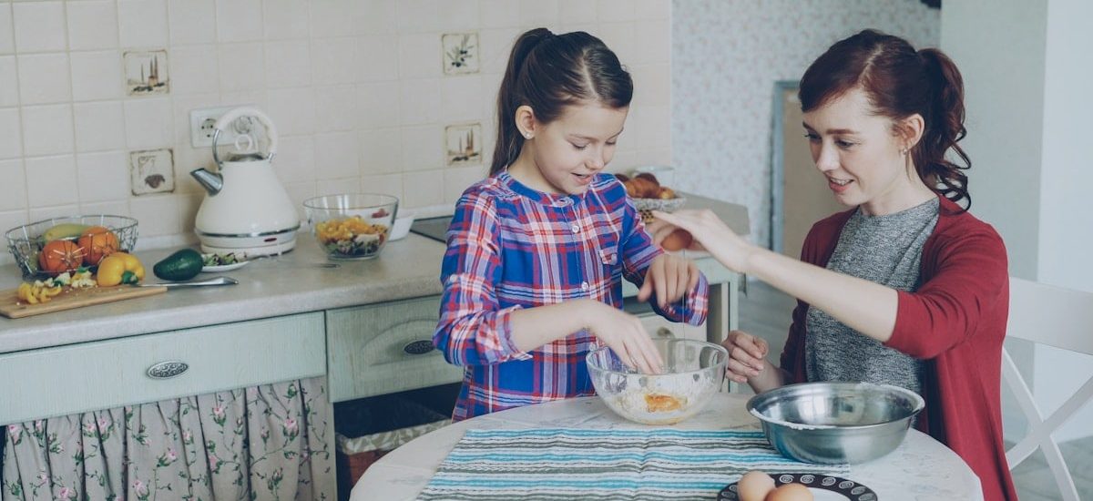 Little cheerful and funny girl helping her mother in the kitchen cropping egg into bowl and mixing dough for cookies. Family, food, home and people concept