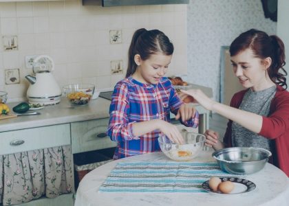 Little cheerful and funny girl helping her mother in the kitchen cropping egg into bowl and mixing dough for cookies. Family, food, home and people concept