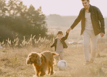 A father and child enjoy a playful outdoor moment with their dog in a grassy field.